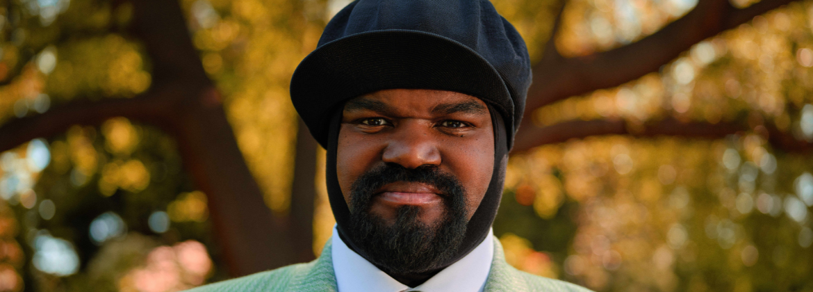 A black man wearing a hat stood in front of the tree smiling at the camera, dressed smart.