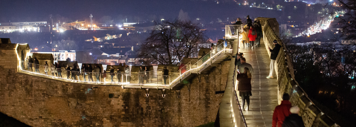 A medieval castle wall illuminated with lights and people walking along the wall walk at night with the city lights in the background