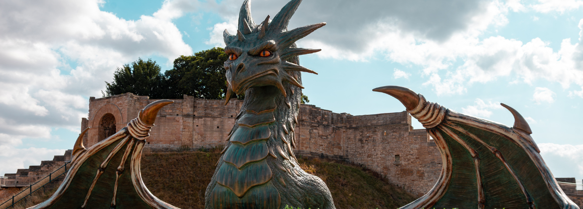 A large dragon sculpture with outstretched wings stands in the foreground at Lincoln Castle. The dragon has glowing orange eyes and detailed scales, with the castle walls visible behind it under a cloudy blue sky.
