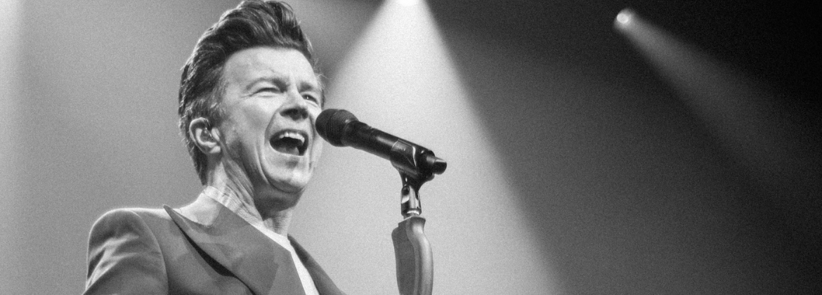 Black and white image of Rick Astley singing into a microphone with spotlights in the background