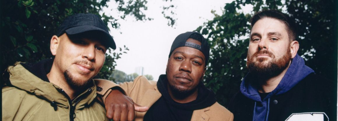 Three members of the band 'Rudimental' pose closely together outdoors, wearing casual jackets, hoodies and caps, with leafy trees in the background.