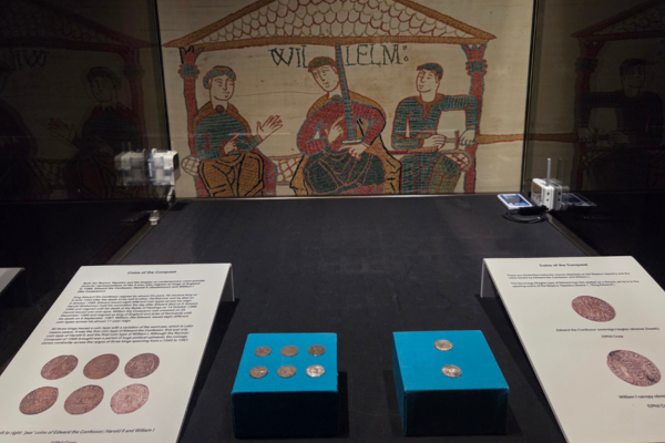 Coins from the Coins of the Conquest exhibition displayed in a case, with a Bayeux Tapestry scene shown behind them.
