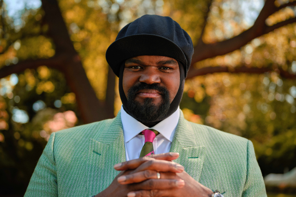 A man with a beard wearing a suit and a hat, stood in front of a tree.